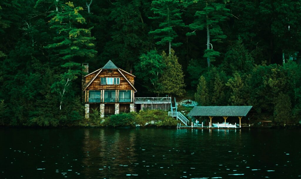 Wooden lakeside cabin with stairs descending to a covered boat dock among tall trees.