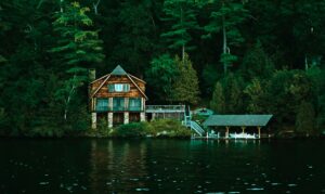 Wooden lakeside cabin with stairs descending to a covered boat dock among tall trees.