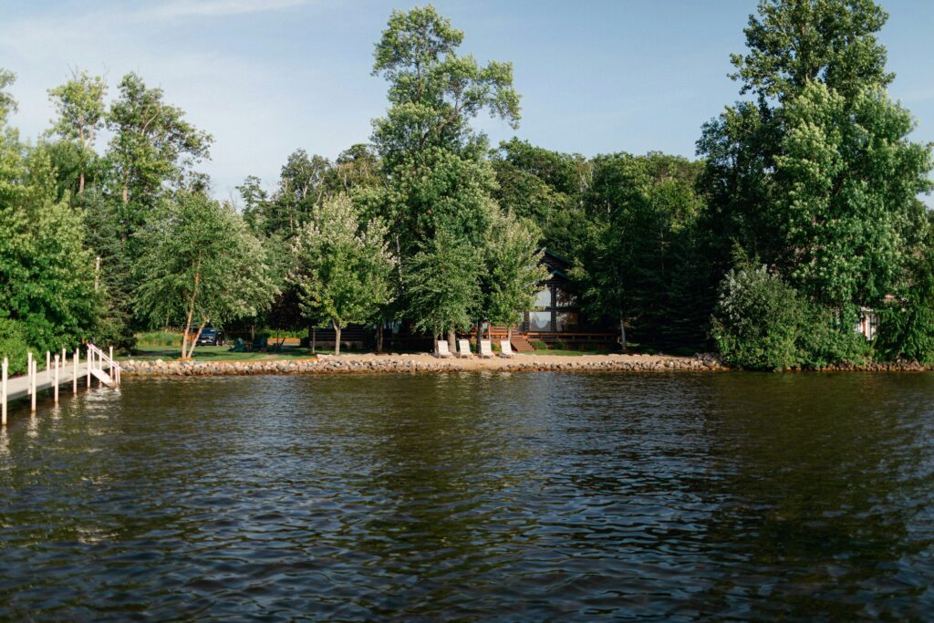 Tree-lined waterfront with a cabin, shoreline chairs, and a dock.